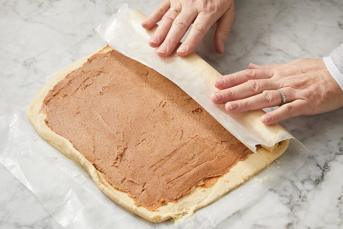 Hands are rolling up dough spread with a brown filling on a marble surface, using wax paper to assist in rolling. The dough is being prepared to make a rolled pastry or dessert.