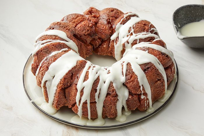 3/4th shot of monkey bread on a white plate; a creamy white icing is drizzled over the top, the plate sits on a light colored countertop with a subtle marble pattern, to the right of the plate, a small, gray bowl contains extra white icing