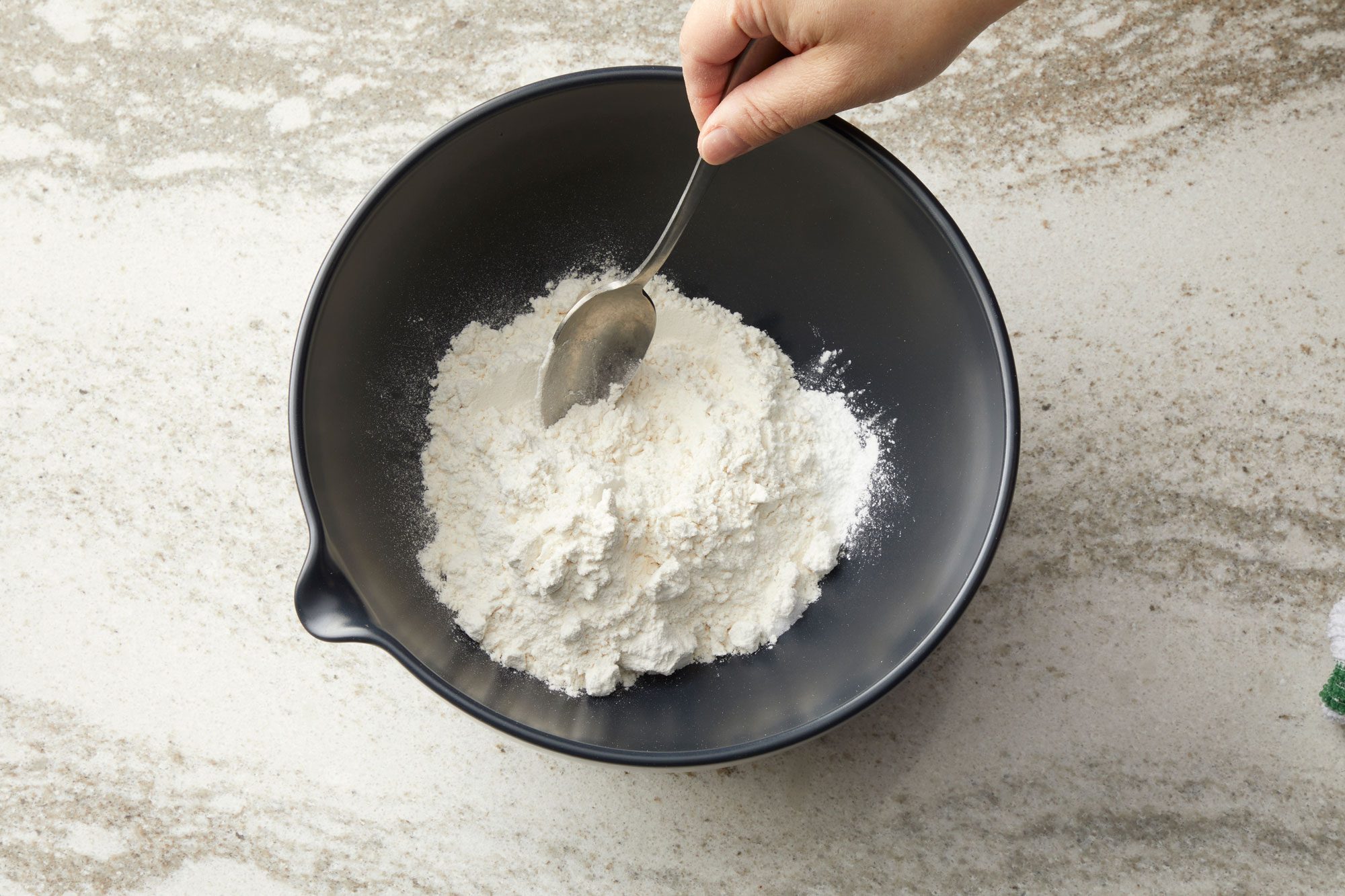overhead shot of a dark gray bowl resting on a light colored, speckled countertop; inside the bowl is a mound of white powder, likely flour, with a silver spoon inserted into it; a hand is visible at the top of the frame, holding the spoon and partially submerging it in the flour; the countertop appears to be a marble or granite surface