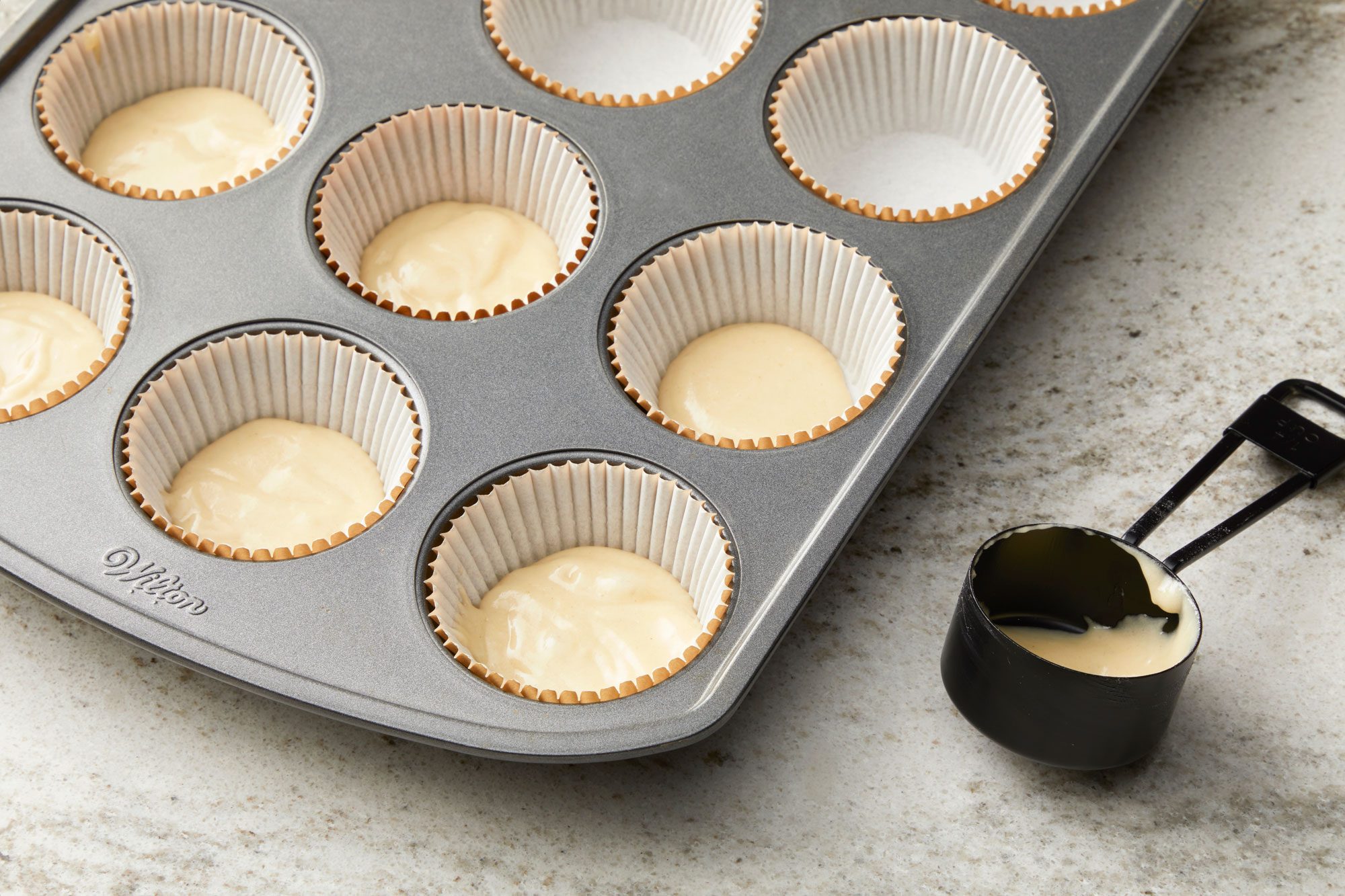 a closeup, top down view of a muffin tin filled with cupcake liners and batter; ten of the depressions are lined with white paper liners, and eight of those liners are filled with a light colored batter; in the lower right corner, a black plastic measuring cup containing some of the same light colored batter is visible; the countertop is a light gray with a subtle speckled pattern