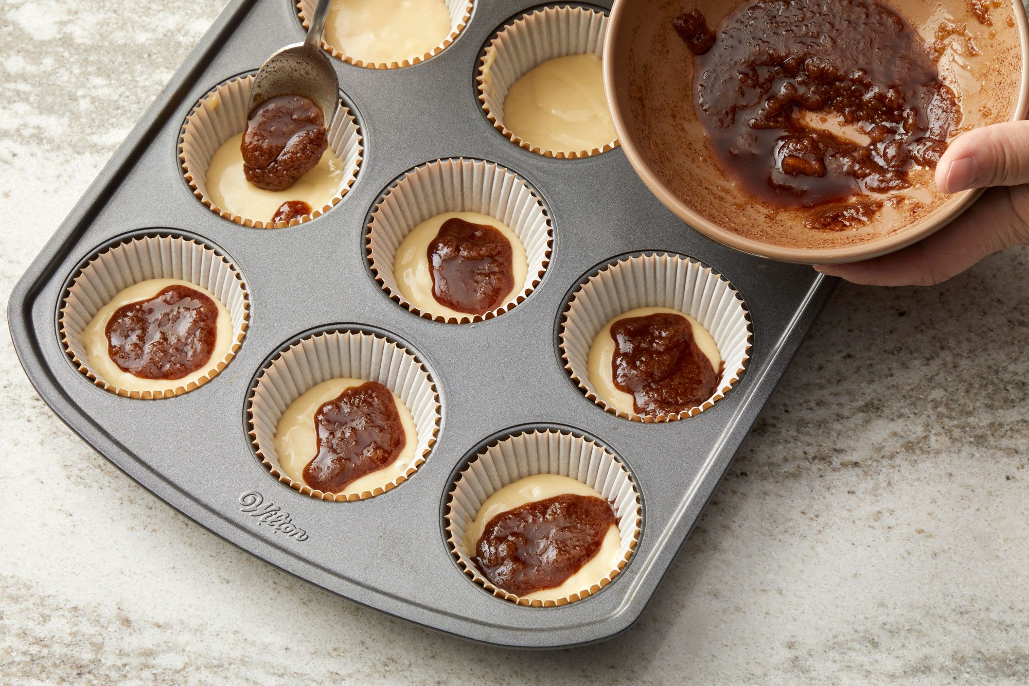 3/4th shot of a muffin tin being prepared for baking; the tin, dark gray and rectangular, contains several white cupcake liners, some of which are filled with a light colored batter; a silver spoon is positioned over one of the batter filled liners, dispensing a dollop of a dark, thick substance, chocolate filling, from its bowl; the countertop beneath the muffin tin is a light gray with a subtle speckled pattern
