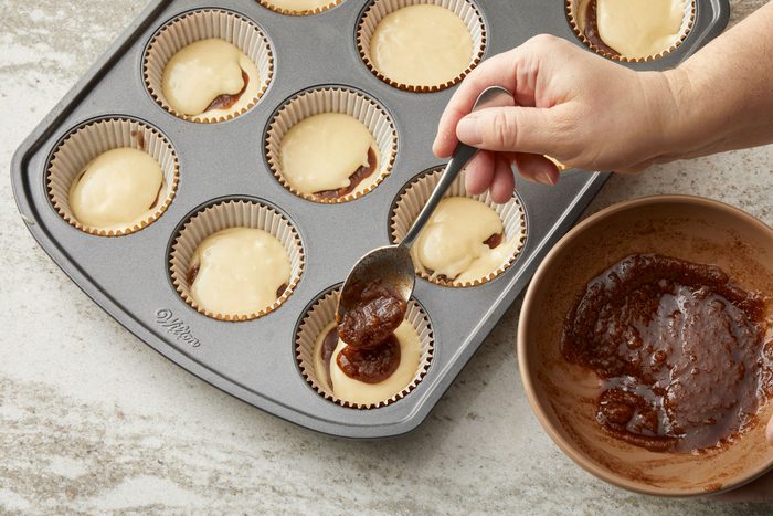 3/4th shot of a gray rectangular muffin tin filled with white paper liners and batter; several liners are already filled with light batter, while a few have a darker substance, likely a jam or fruit filling, added on top; a hand is visible in the right side of the frame, holding a silver spoon and adding more of the dark filling from a tan bowl; the bowl, partially visible, contains the thick, dark substance; the countertop is a light gray with a subtle speckled pattern