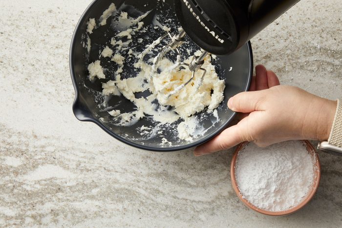 overhead shot of a dark gray bowl on a light colored, speckled countertop; inside the bowl, a white, creamy substance, likely cream cheese or frosting, is being mixed with a black KitchenAid hand mixer; partially submerged in the white mixture; a hand is also visible, to the right of the bowl, a smaller, tan bowl contains a white powder, possibly flour or powdered sugar; the countertop appears to be a marble or granite surface
