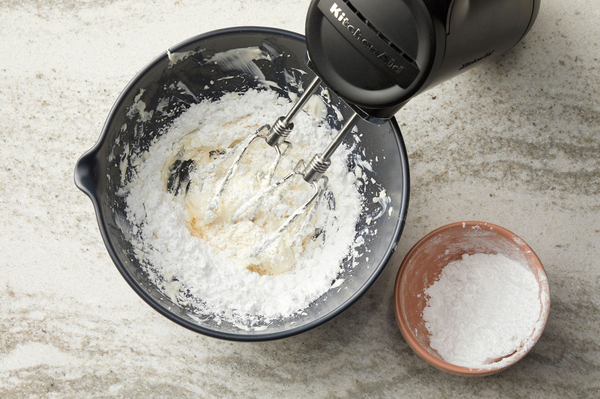 overhead shot of a dark gray bowl on a light colored, speckled countertop; inside the bowl, a white, creamy substance, likely cream cheese or frosting, is being mixed with a black KitchenAid hand mixer; partially submerged in the white mixture; to the right of the bowl, a smaller, tan bowl contains a white powder, possibly flour or powdered sugar; the countertop appears to be a marble or granite surface