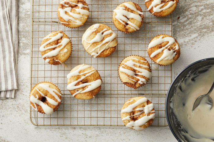 overhead shot of a batch of small, oval shaped baked cinnamon rolls, arranged on a gold wire cooling rack; drizzled with a white icing; in the lower right corner, a dark bowl containing leftover icing and a spoon is visible; the cooling rack sits on a light colored, speckled countertop