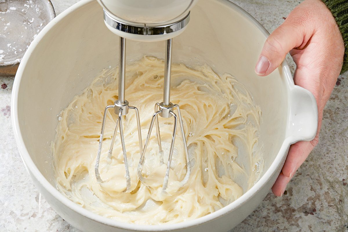 A hand holds a white mixing bowl with creamy batter being mixed by an electric hand mixer. The mixer's metal beaters are partially submerged in the batter. The background shows a gray, textured surface.