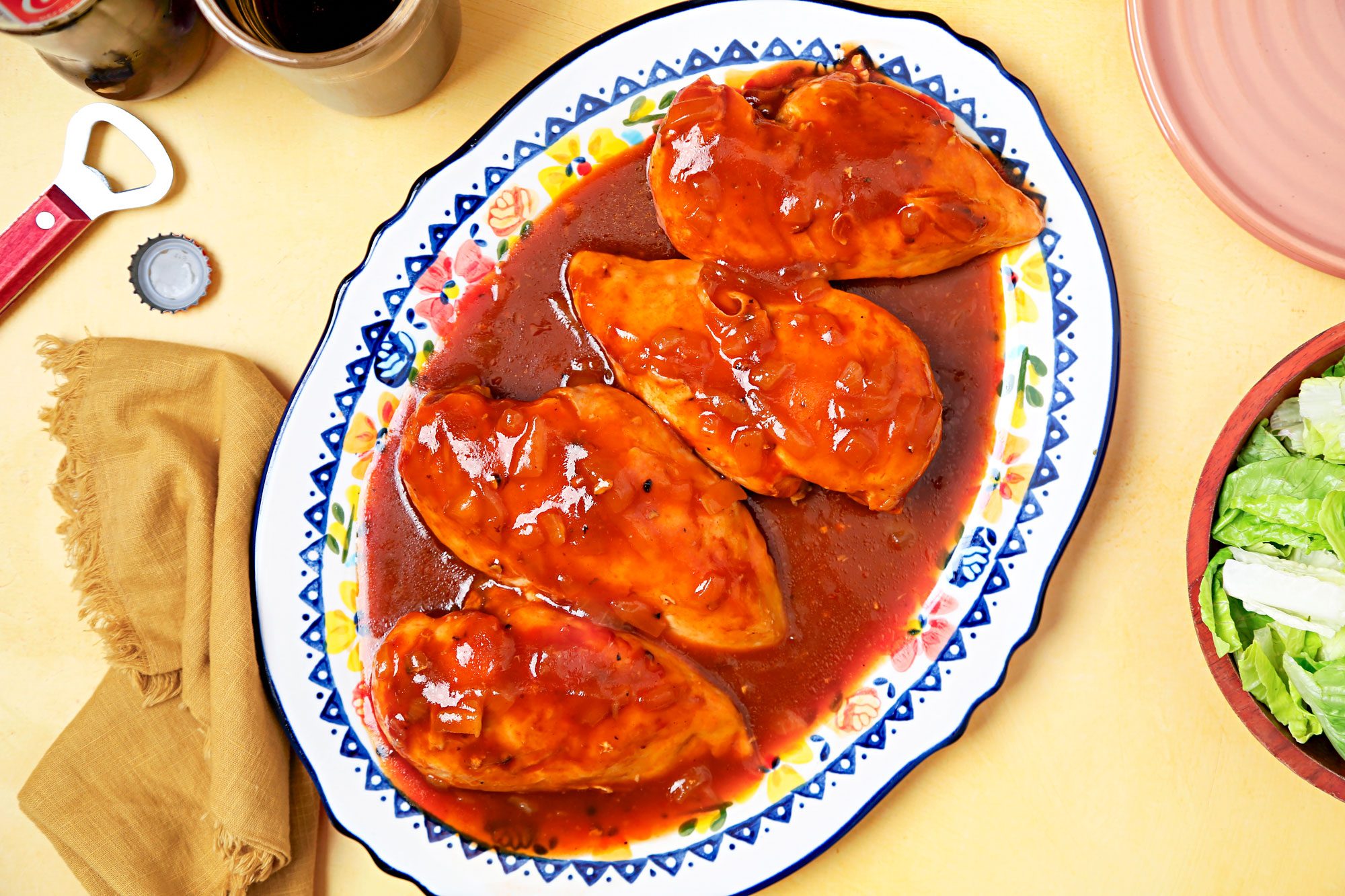 Overhead shot of a dish of Cola Chicken, this particular dish is displayed on a colorful plate with a blue and white pattern and floral accents; there are four chicken breasts covered in a sweet, savory cola sauce; there is a side salad of lettuce in a wooden bowl, and a bottle opener and bottle cap on the table beside the dish; the background is a yellow tablecloth