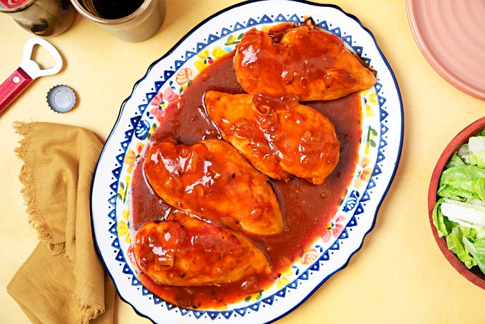 Overhead shot of a dish of Cola Chicken, this particular dish is displayed on a colorful plate with a blue and white pattern and floral accents; there are four chicken breasts covered in a sweet, savory cola sauce; there is a side salad of lettuce in a wooden bowl, and a bottle opener and bottle cap on the table beside the dish; the background is a yellow tablecloth
