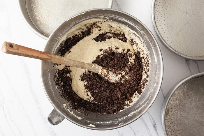 Baking ingredients in a metal bowl, including flour and crushed chocolate cookies, with a wooden spoon stirring them. The bowl is set on a white marble surface with several other empty bowls nearby.