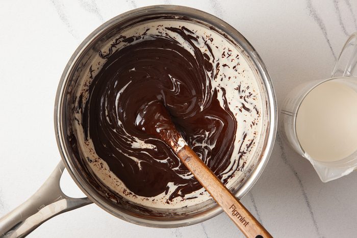 A saucepan with melted chocolate being stirred with a wooden spatula, creating a swirled pattern. A small pitcher of milk is placed beside the saucepan on a white marble surface.
