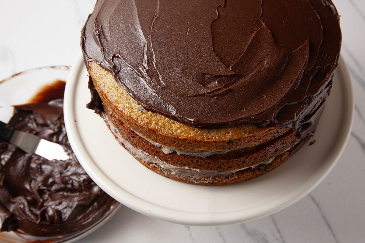 A layered chocolate cake with dark frosting sits on a white cake stand. A bowl with chocolate frosting and a spatula is beside the cake on a marble surface.