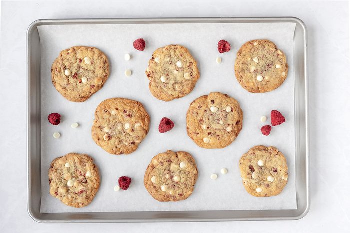 A baking tray with nine chocolate chip cookies and scattered raspberries. The cookies are arranged on parchment paper, showing a golden-brown color with visible white chocolate chips.