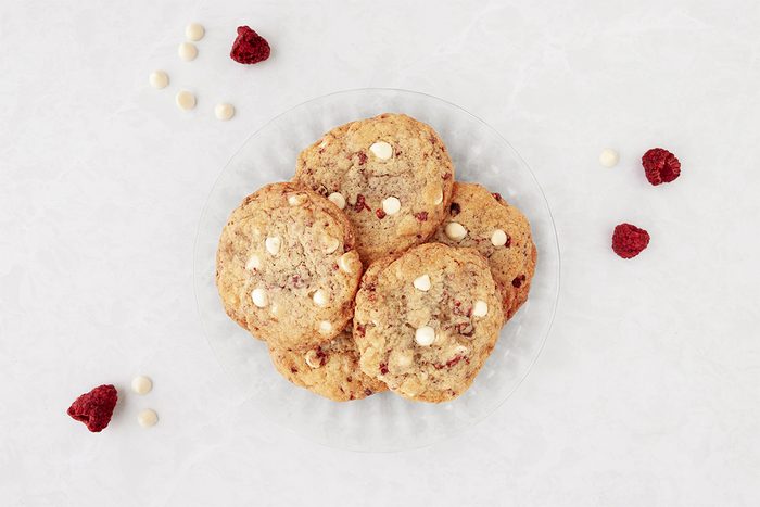 A plate of cookies with white chocolate chips, surrounded by scattered raspberries and white chocolate chips on a light surface.
