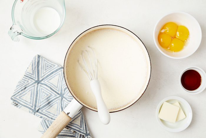 A mixing bowl with creamy batter and a whisk is surrounded by a measuring cup of milk, a bowl with three egg yolks, a small bowl of vanilla extract, a dish of butter, and a blue patterned cloth on a white surface.
