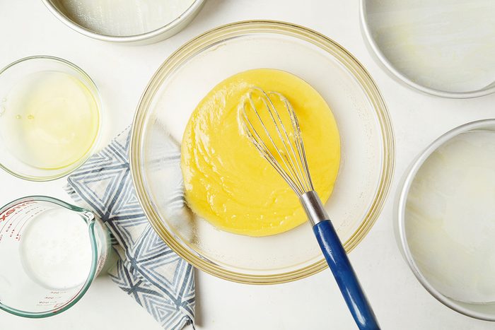 A glass bowl filled with yellow cake batter and a blue whisk on a white surface. Surrounding items include empty round baking pans, a textured gray cloth, and a measuring cup with a small amount of liquid.