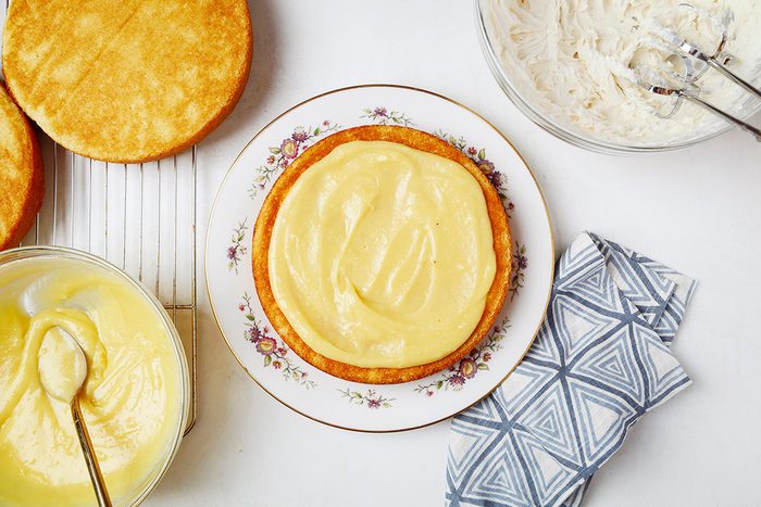 A round cake base is being frosted with yellow icing on a decorative plate. Nearby, there's a bowl of more yellow icing with a spoon, a bowl with a hand mixer, and a folded patterned cloth. Another cake layer is on a cooling rack.
