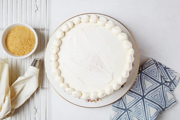 A round white cake with smooth icing and decorative piping along the edge, placed on a white plate. Nearby are a bowl of light brown sugar, a piping bag, and a blue patterned napkin on a light surface.