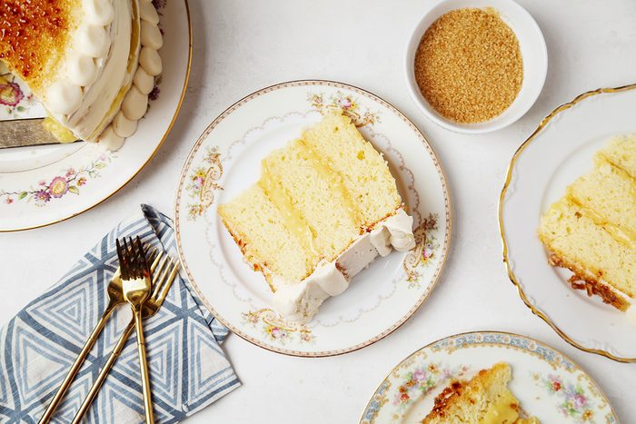 Slices of layered vanilla cake with frosting on elegant floral plates, accompanied by a bowl of brown sugar. A fork and knife rest on a patterned napkin nearby.