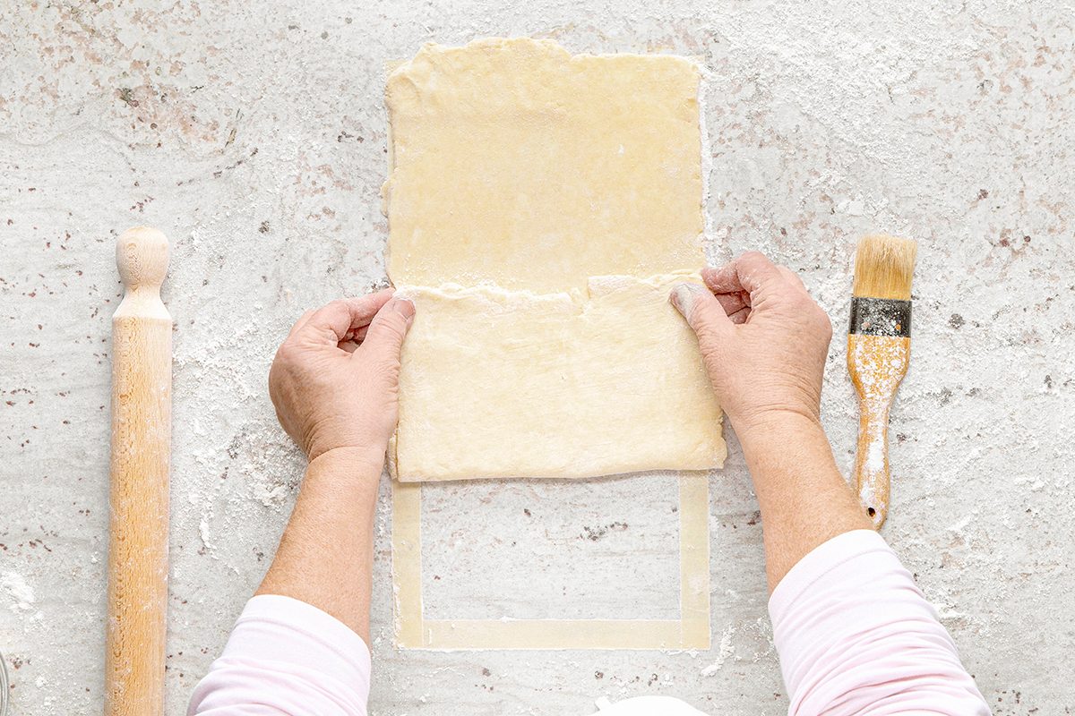 Hands folding a sheet of dough on a floured surface with a rolling pin and a pastry brush nearby.