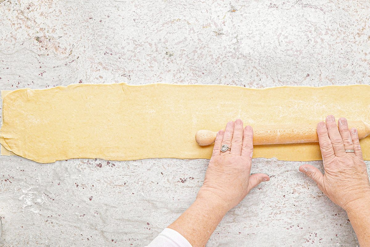 Hands rolling out a sheet of dough with a wooden rolling pin on a lightly floured surface. The dough is thin and rectangular, and the person is wearing a white long-sleeved shirt.