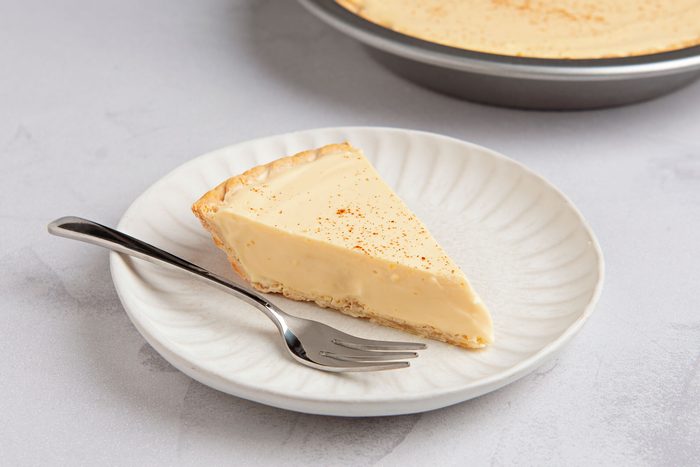closeup shot of a slice of custard pie on a white plate with a fork next to it; the pie is dusted with cinnamon