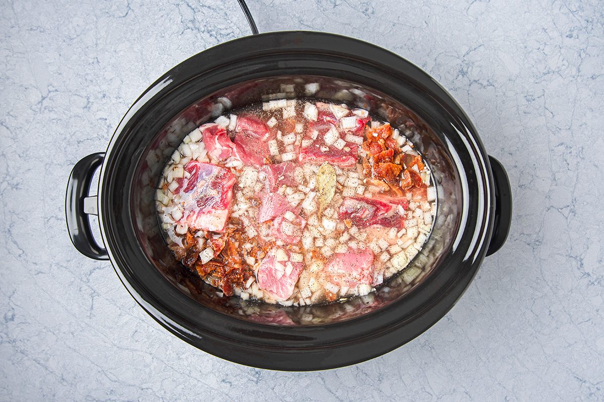 Overhead view of a slow cooker with uncooked ingredients, including chunks of raw meat, chopped onions, and spices mixed with broth. The slow cooker sits on a marble countertop.