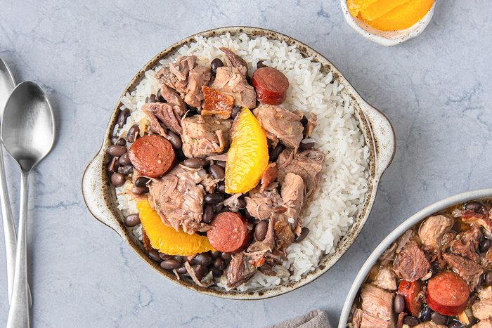 A bowl of rice topped with black beans, slices of sausage, chunks of pork, and orange segments, served on a speckled plate.