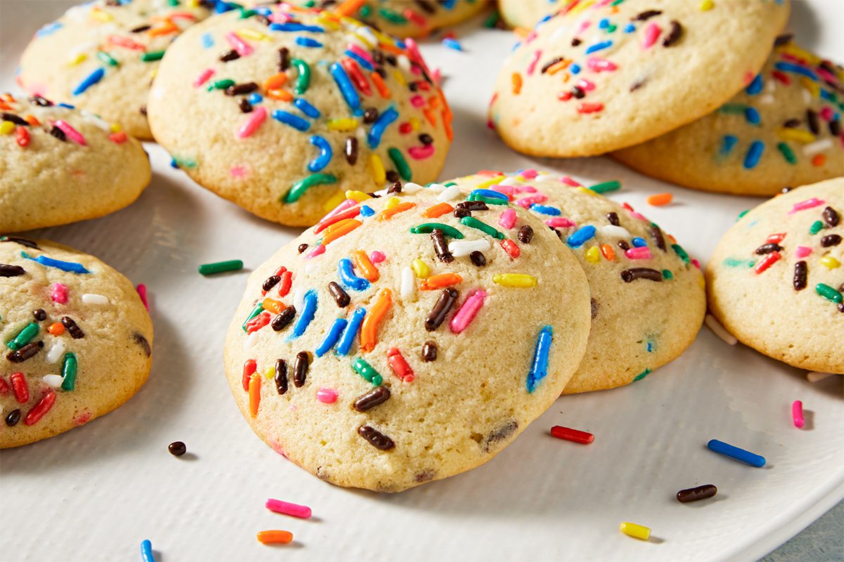 A plate of round, soft cookies topped with colorful sprinkles. The cookies are light golden brown, scattered with red, green, yellow, blue, and chocolate sprinkles, set against a simple, light-colored background.