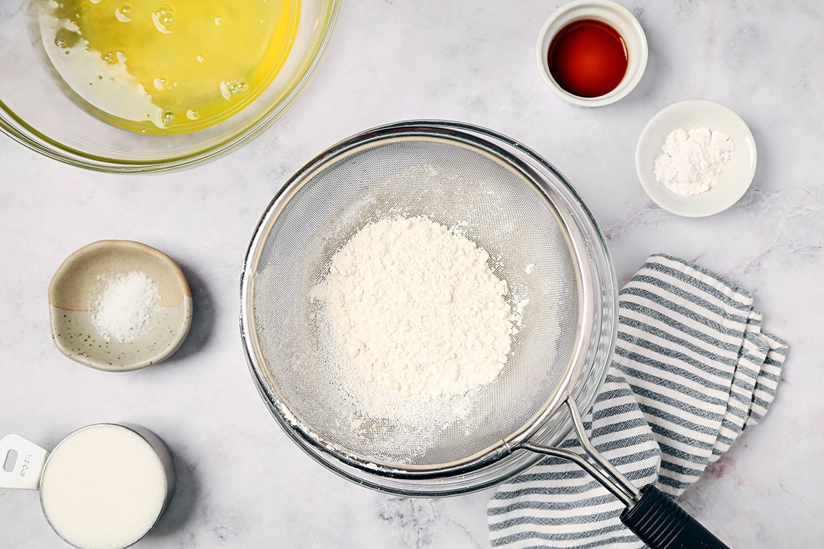 Baking ingredients on a countertop include a sieve with flour, a small bowl of vanilla extract, baking powder, salt, milk in a measuring cup, and a bowl of egg whites. A striped cloth lies nearby.