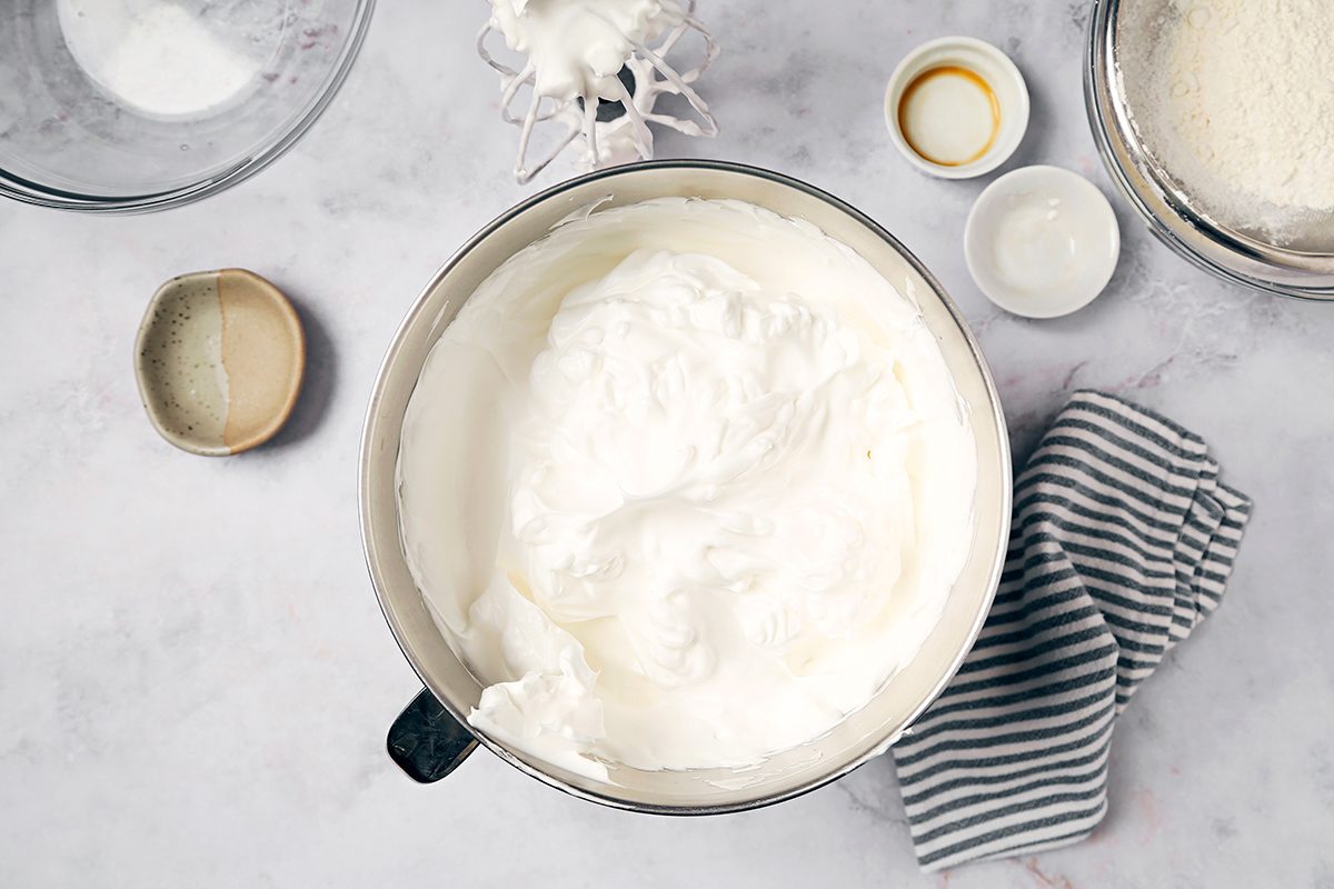 A mixing bowl filled with whipped cream on a countertop. Surrounding it are ingredients in small bowls, including sugar and vanilla extract. A striped cloth lies nearby, and a sieve with flour is partially visible.