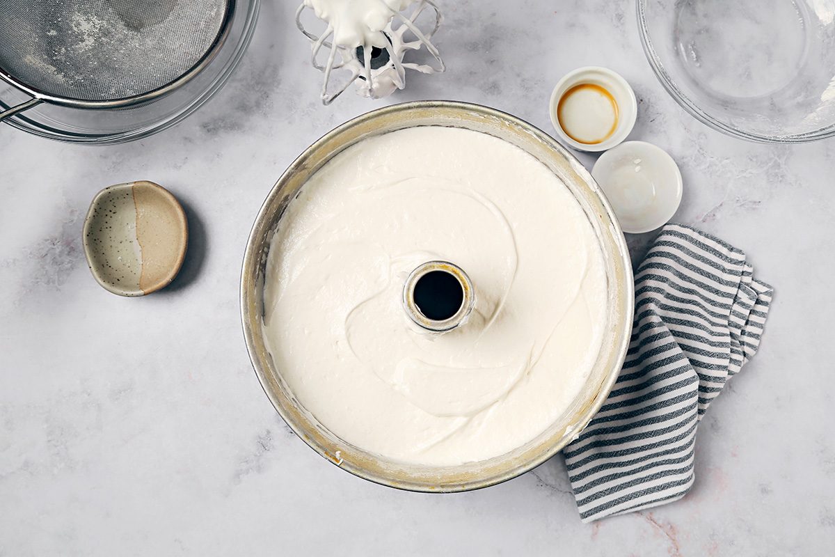 A tube pan filled with cake batter on a countertop, surrounded by a striped cloth, a fine-mesh strainer, and small bowls containing different ingredients.