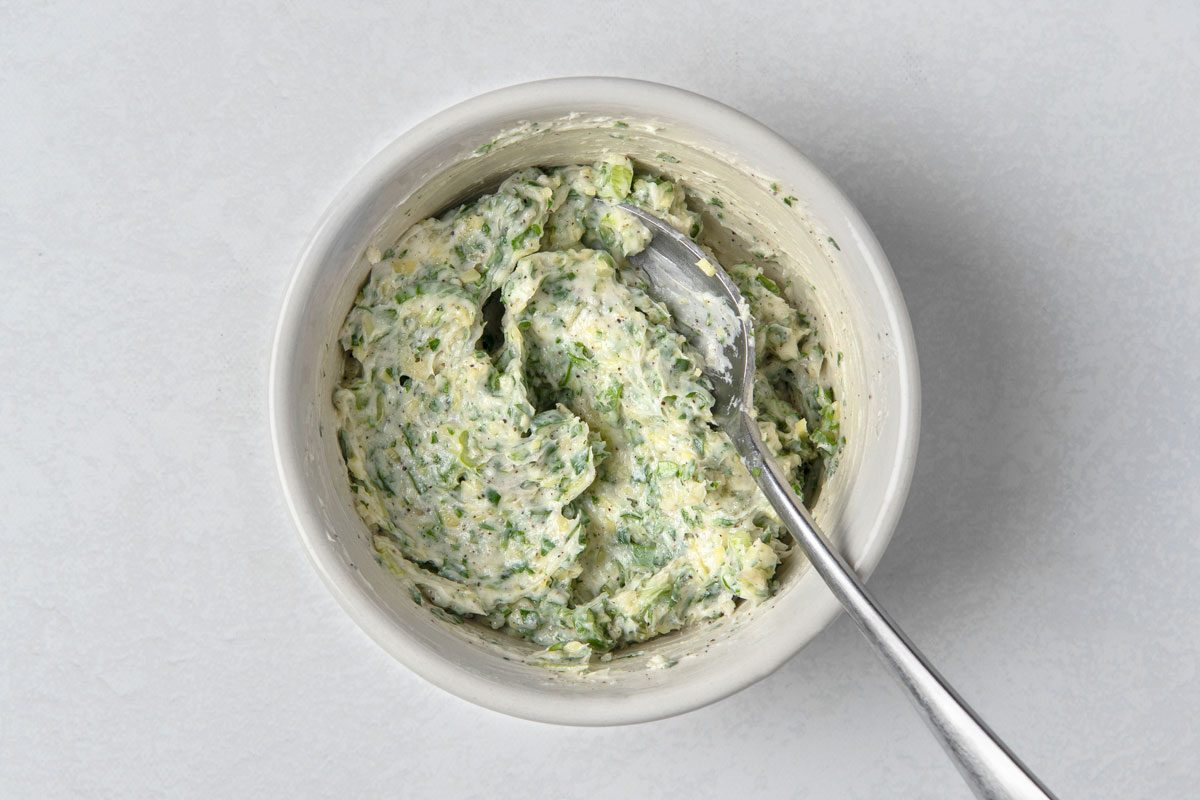 overhead shot of a small white bowl filled with green herb butter; a silver teaspoon is also in the bowl; the bowl and spoon are on a white surface