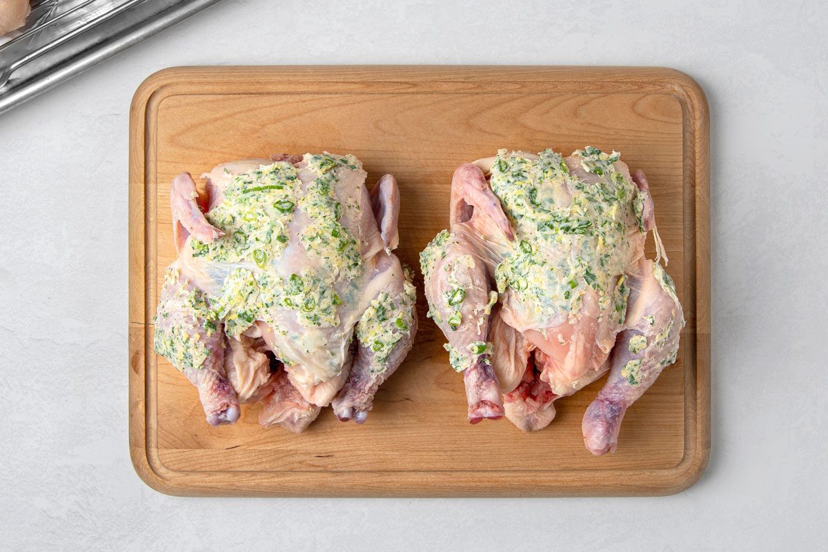 overhead shot of two raw whole chickens on a wooden cutting board; the chickens are coated in a green herb butter mixture; the cutting board is on a white countertop; in the upper left corner of the image, a metal baking rack with foil lining is partially visible