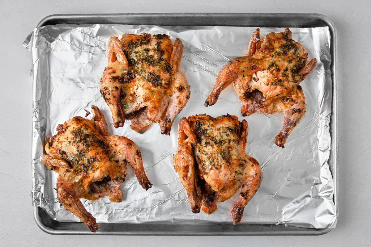 overhead shot of four roasted chickens on a baking sheet lined with aluminum foil; the chickens are covered in a layer of herbs and spices