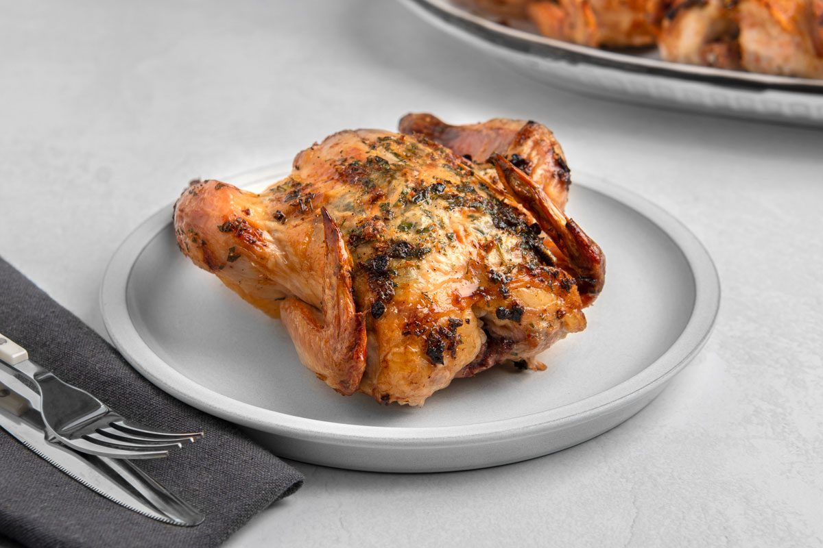 closeup shot of a roasted chicken, sitting on a white plate and the plate is on a white table; to the left of the plate is a black napkin with a fork and knife on it; there is a blur in the background which looks like another roasted chicken on a plate