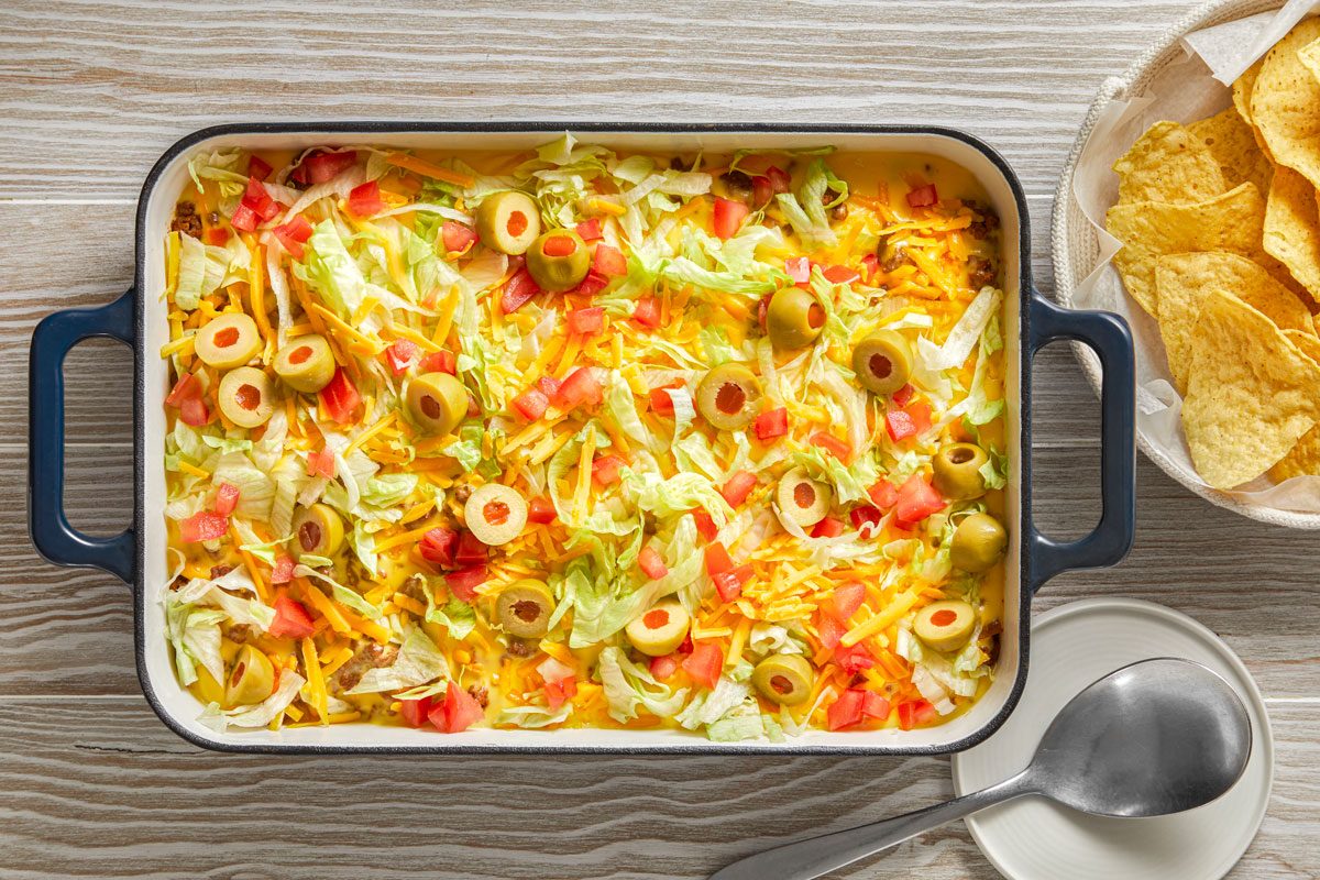 Top view shot of Haystack Supper; In a baking pan; top with lettuce; cheddar cheese; tomatoes and olives; serve with chips; serving spoon; wooden surface;