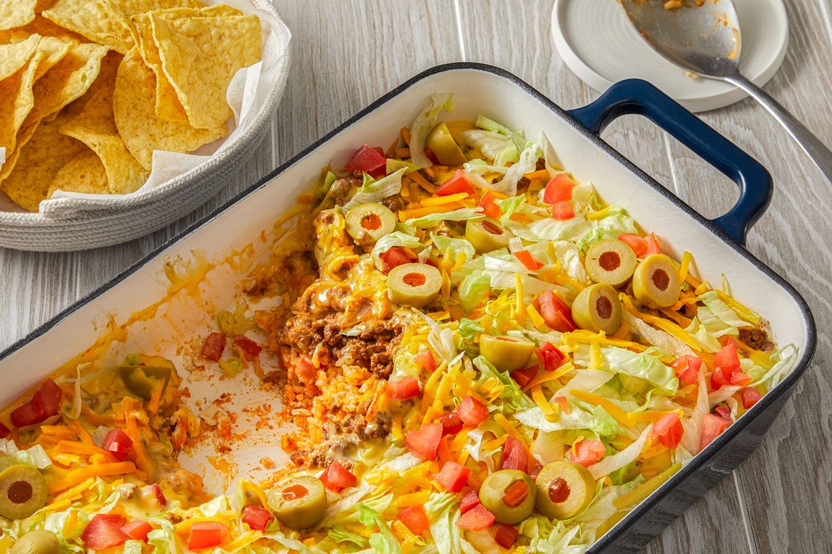 Table view shot of Haystack Supper; In a baking pan; top with lettuce; cheddar cheese; tomatoes and olives; serve with chips; serving spoon; wooden surface;
