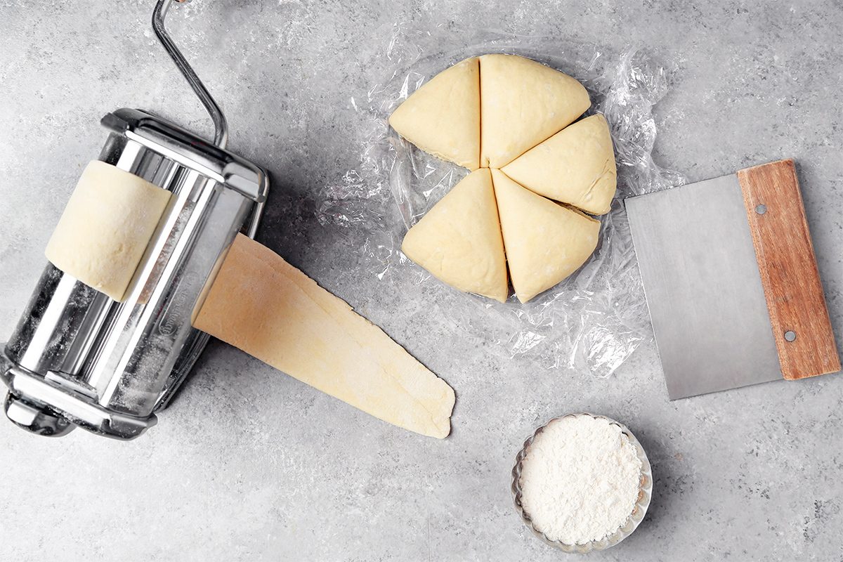 A pasta maker with dough being rolled out, beside a circle of dough divided into six triangles. A bowl of flour and a metal dough scraper are also on the gray countertop.