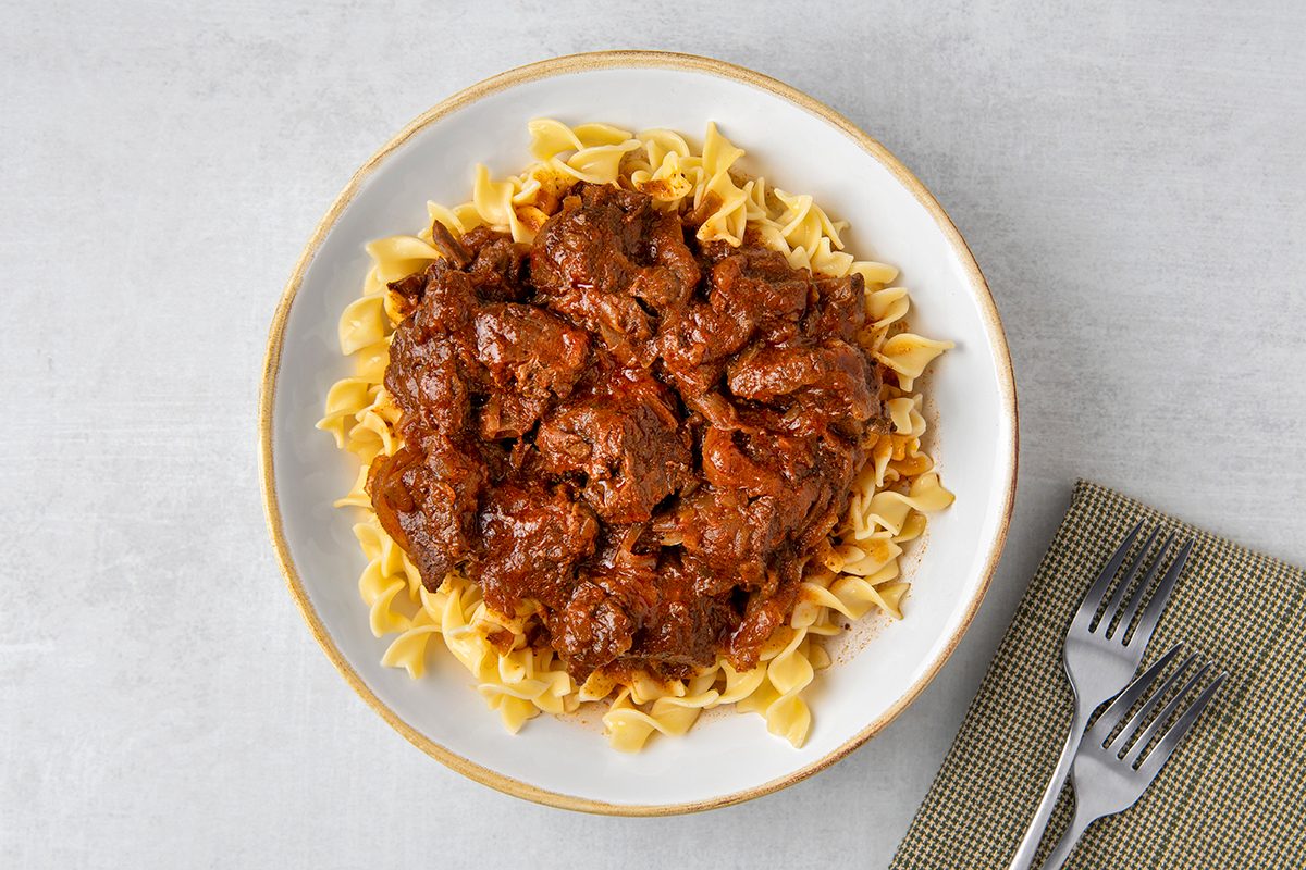 A plate of beef stew served over a bed of egg noodles. The dish is placed on a light gray surface next to a folded beige cloth with two forks.