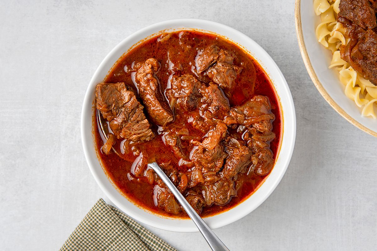 A bowl filled with rich, red beef stew containing chunks of meat and onions. A spoon is resting inside the bowl. The stew is accompanied by a beige cloth napkin on the left and a partial view of pasta on the right.
