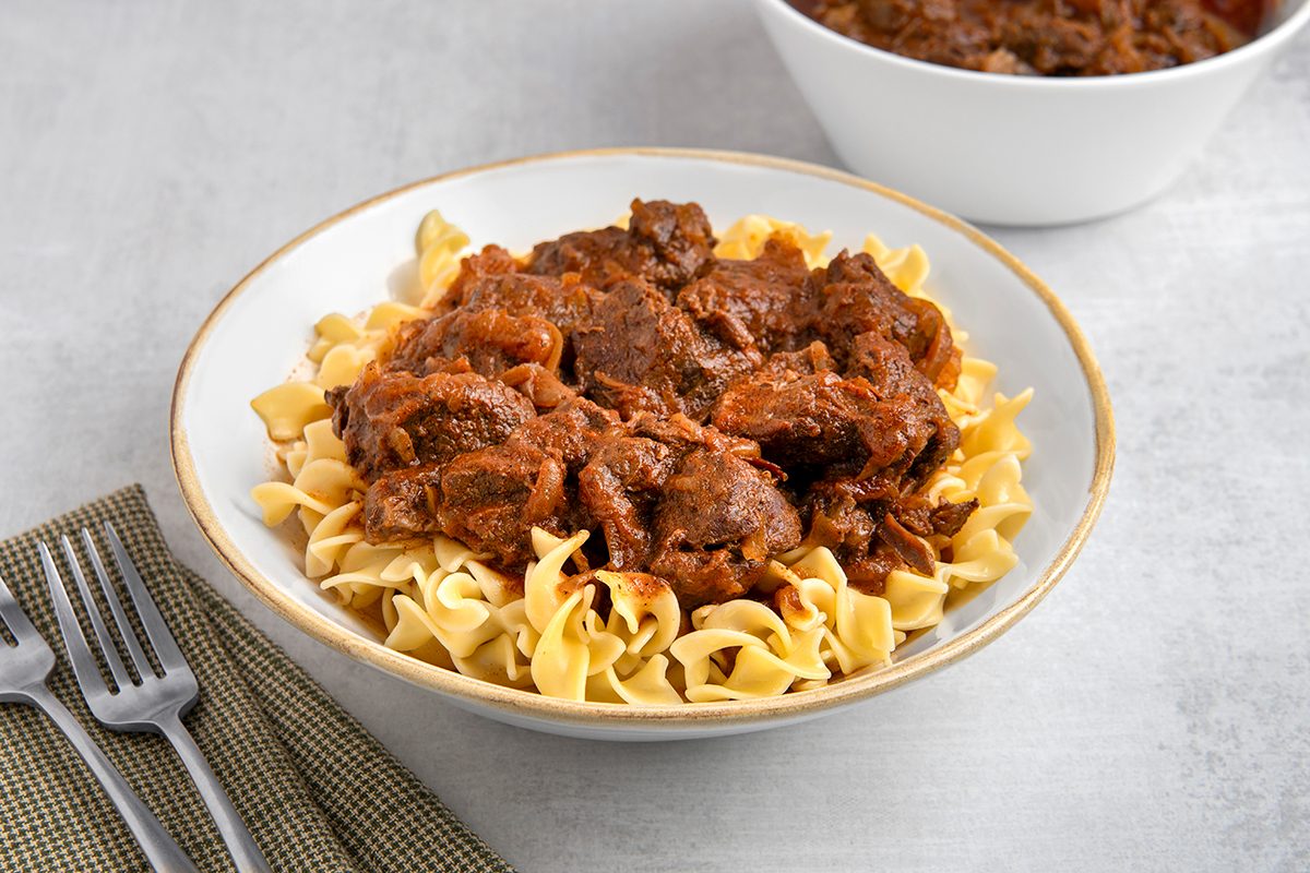 A bowl of beef goulash served over wide egg noodles, placed on a light-colored surface. A textured napkin with two forks is on the left. In the background, there's a white bowl with more goulash.