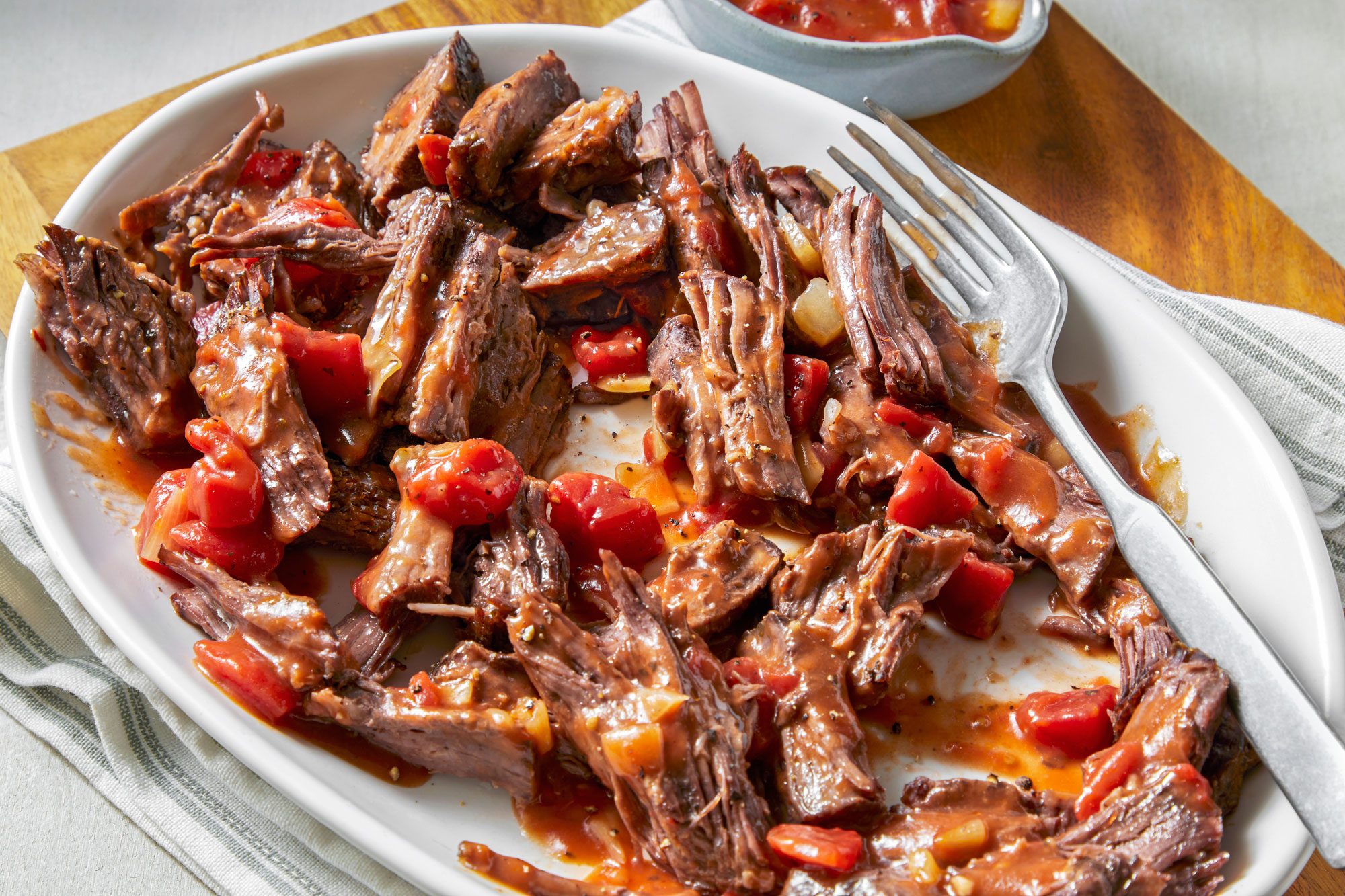 3/4th shot of Italian pot roast, presented on a white oval platter; a silver fork rests beside the roast, and in the background, a small bowl of sauce can be seen; the plate is set on a wooden surface