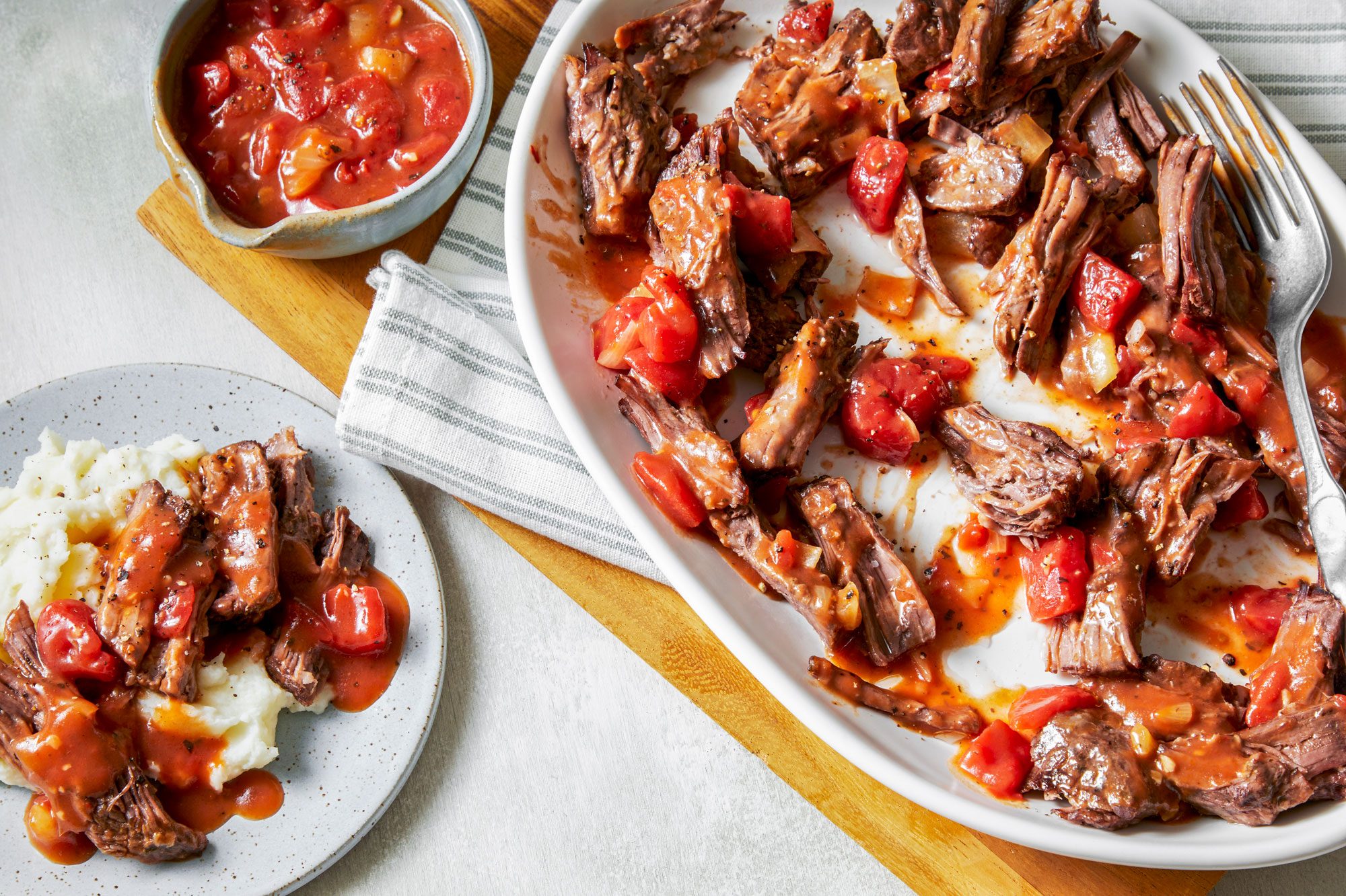 overhead shot of Italian Pot Roast served in a large white oval dish; on side plate mashed potatoes topped with additional gravy and some of the meat; next to it, there’s a small bowl of a tomato based sauce; the background is a light colored table surface and a striped cloth