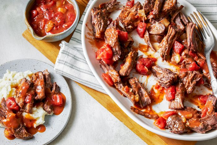 overhead shot of Italian Pot Roast served in a large white oval dish; on side plate mashed potatoes topped with additional gravy and some of the meat; next to it, there’s a small bowl of a tomato based sauce; the background is a light colored table surface and a striped cloth