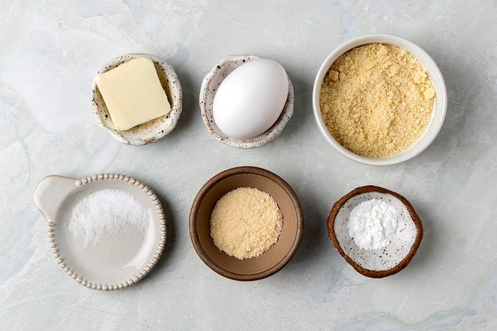 Ingredients arranged on a light surface: a pat of butter, a whole egg, a bowl of almond flour, a dish of baking soda, a small bowl of coconut flour, and a bowl of salt.