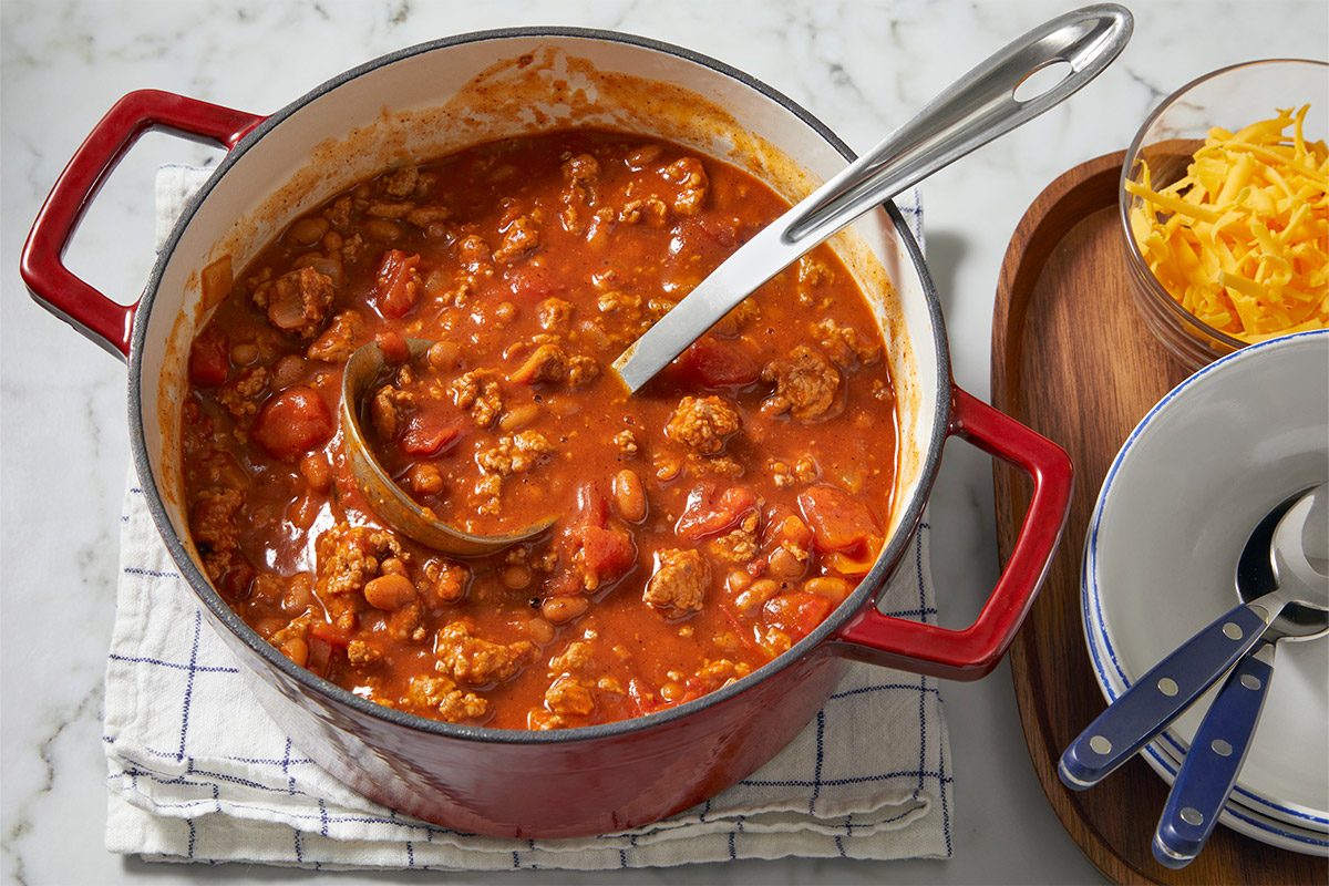 overhead shot of a pot of Kid’s Favorite Chili; the chili is rich and thick, filled with ground meat, beans, and chunks of tomatoes; a metal ladle rests in the pot, a small bowl holds a mound of shredded cheese, while a clean white bowl and spoon are also there; the chili pot is set on a plaid cloth, and the background features a marble countertop
