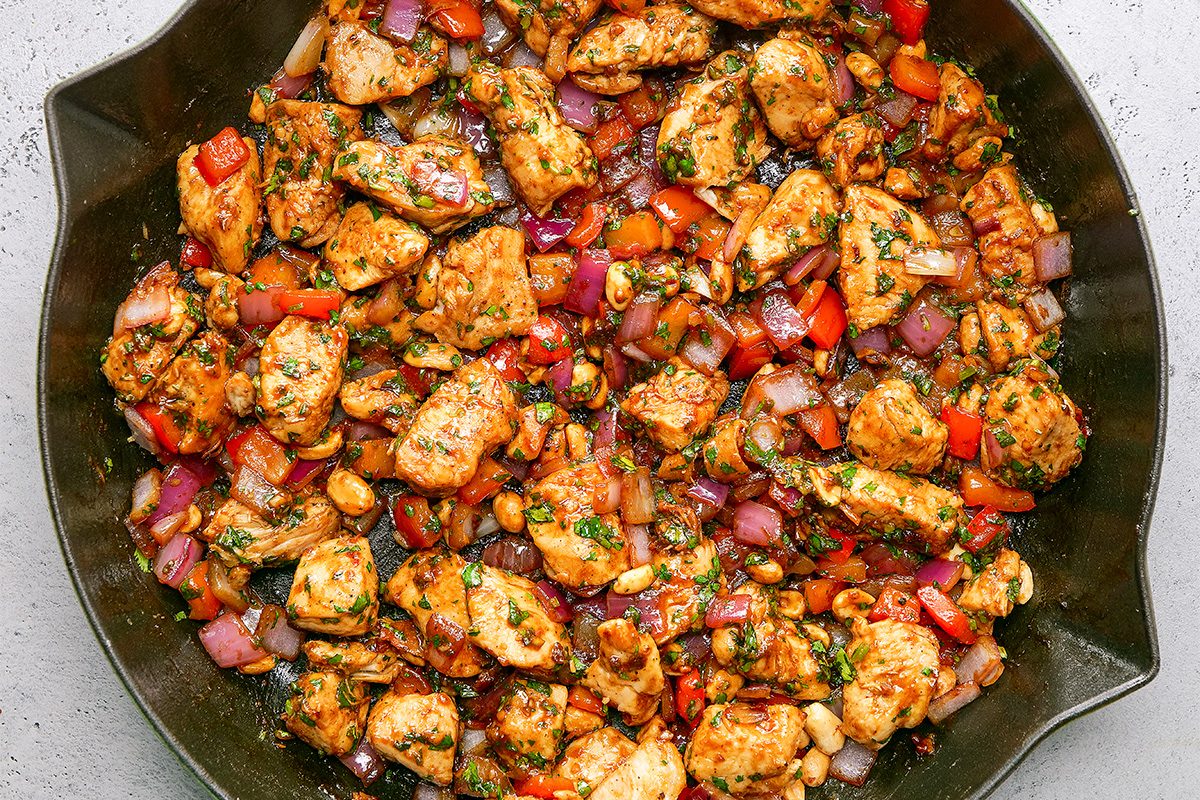A skillet filled with cooked, seasoned chicken pieces mixed with diced red onions, tomatoes, and herbs. The dish has a colorful and appetizing appearance.