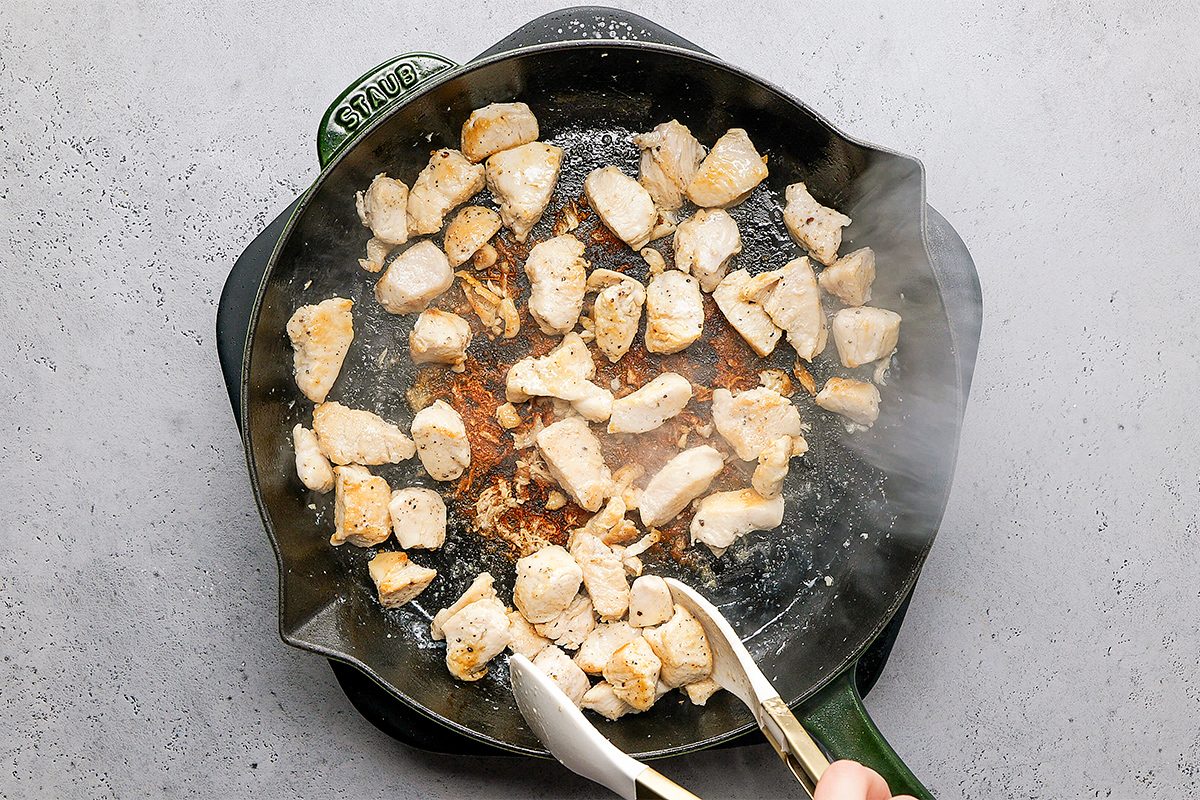 Chunks of chicken being cooked in a black skillet on a stovetop, with some browning visible. A pair of tongs is used to stir or flip the pieces. Steam rises, indicating heat.