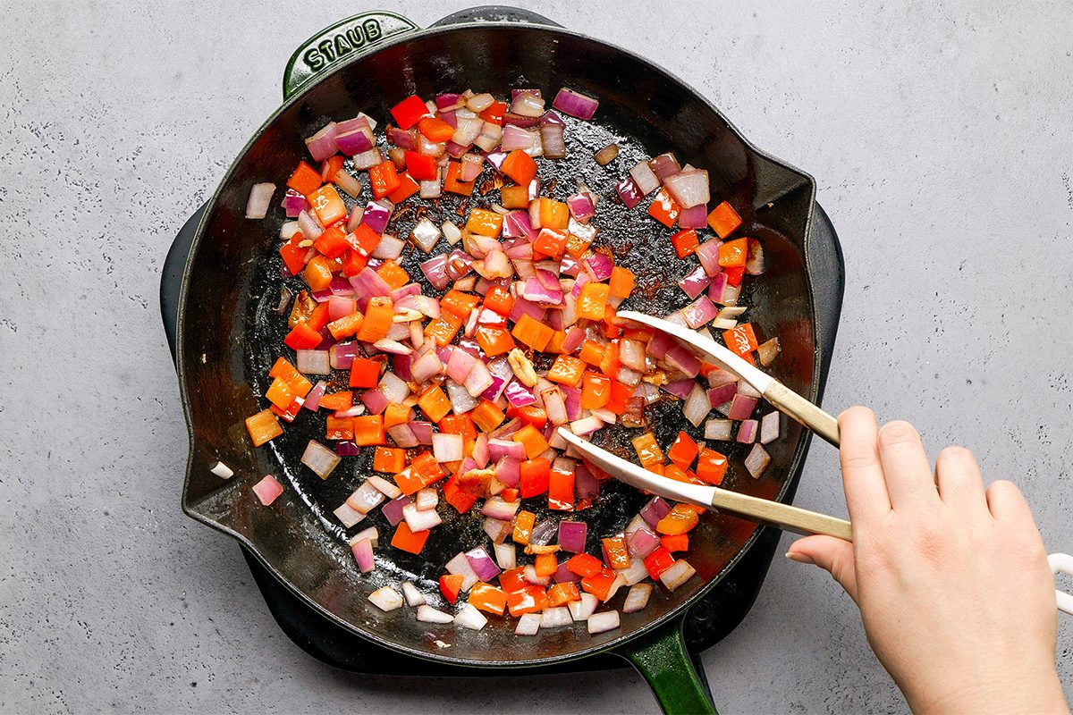 A hand uses tongs to stir diced red onions and red bell peppers in a black skillet. The skillet sits on a green-handled pan over a grey surface.