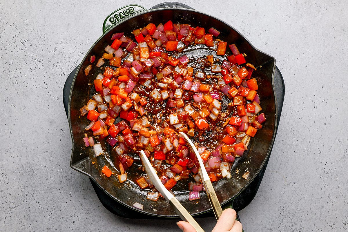 A hand using tongs to stir a mixture of diced red onions, tomatoes, and other vegetables in a cast iron skillet on a gray surface.